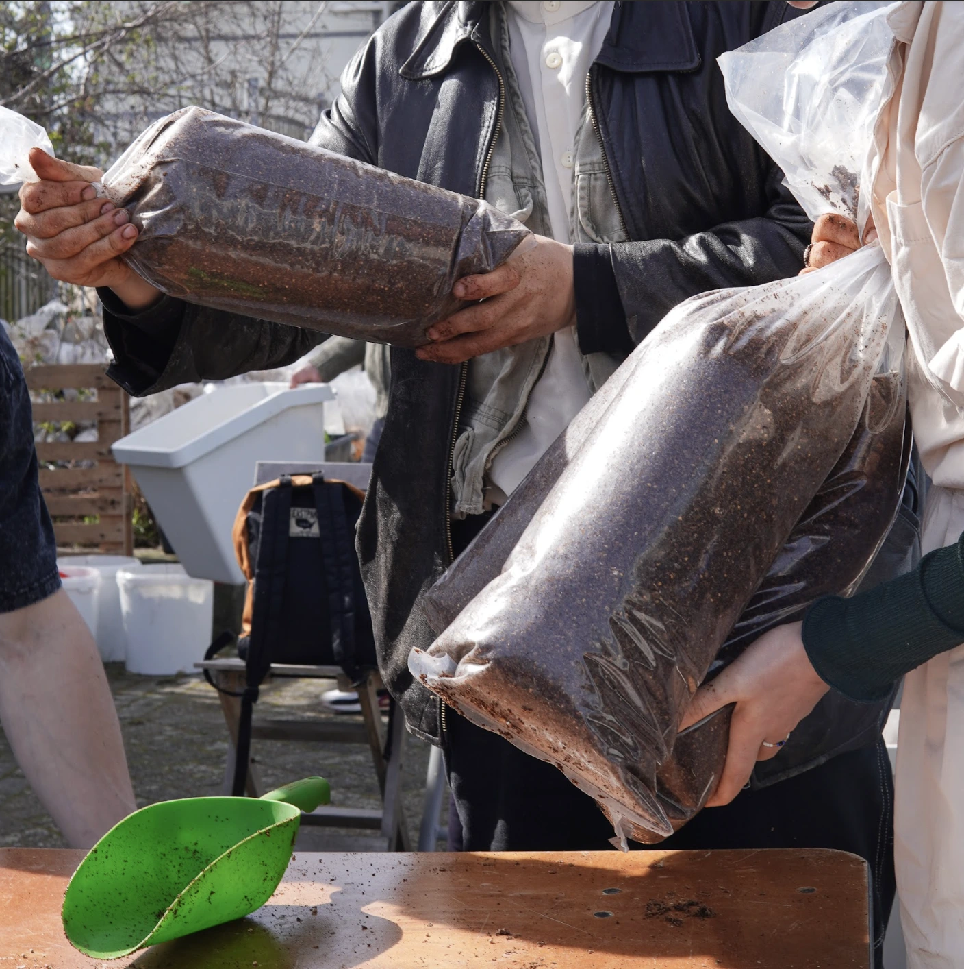 People holding plastic bags filled with coffee grounds near a green garden scoop on a wooden table.