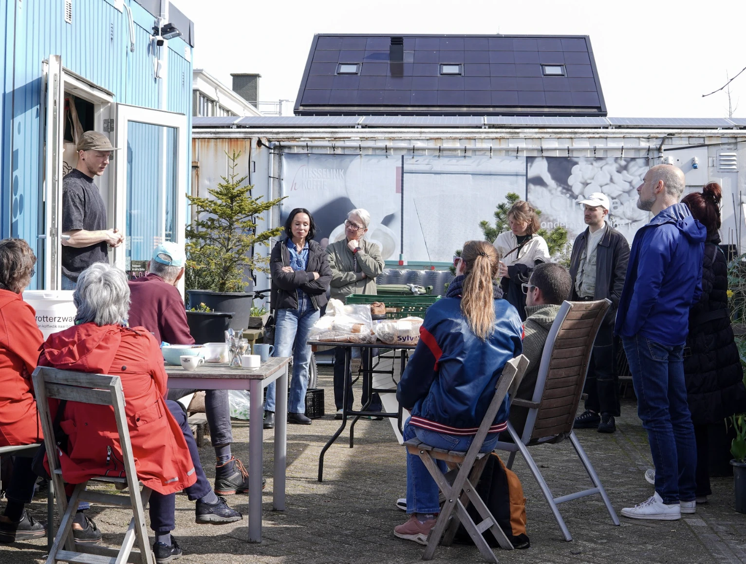 Group of people outdoors listening to a man speaking by a blue building with solar panels in the background.