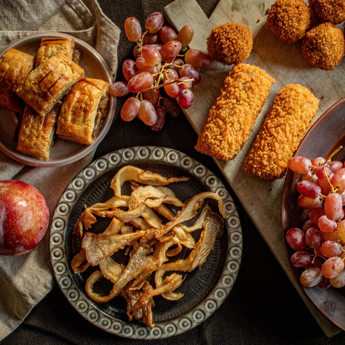 Plates with fried mushrooms, sausage rolls, breaded croquettes, and clusters of red grapes on a dark surface.