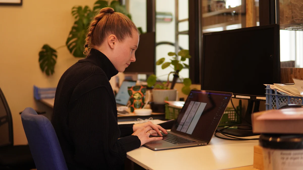 Woman with braided hair typing on a laptop at a desk in a modern office.