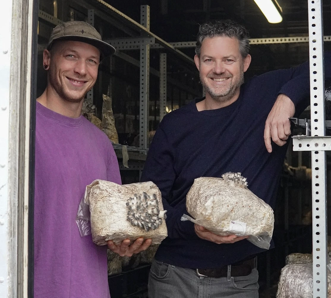 Two men smiling and holding mushroom growing bags inside a mushroom farm.