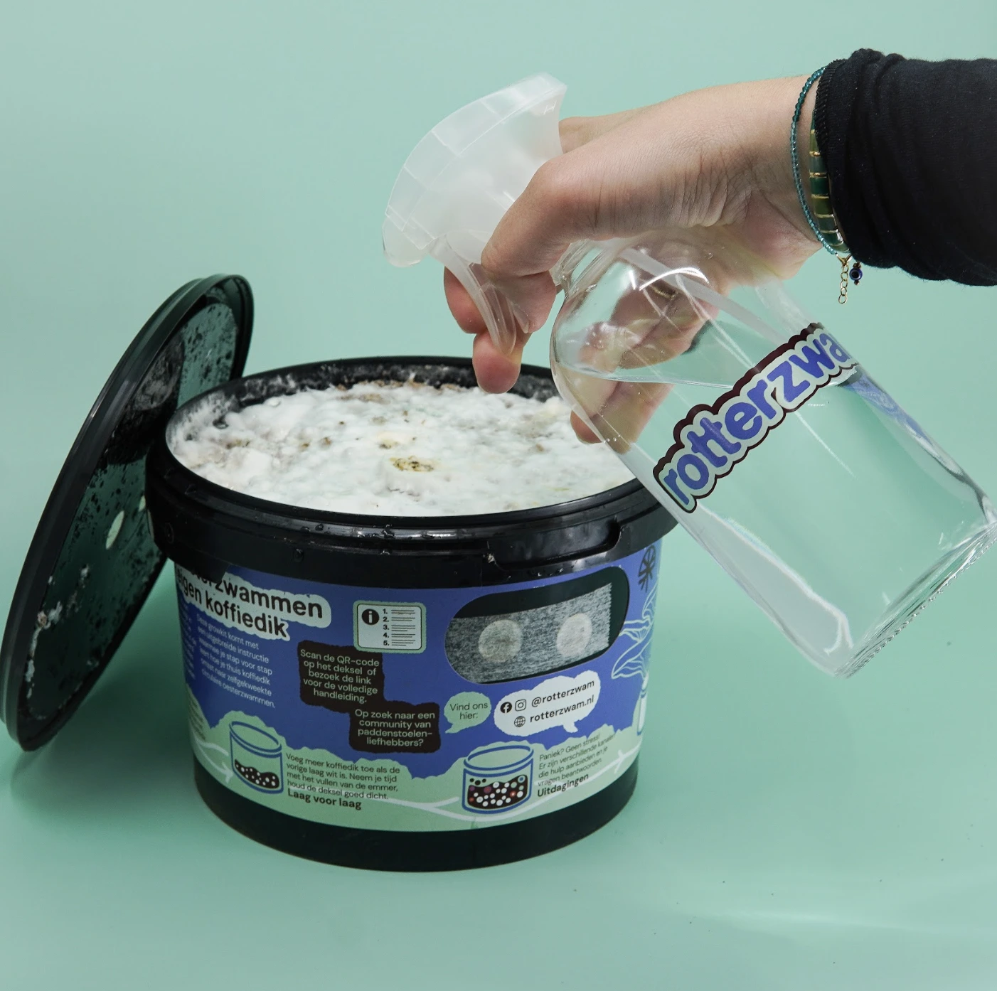 Hand spraying water from a bottle onto a white substance inside a labeled black container with an open lid.