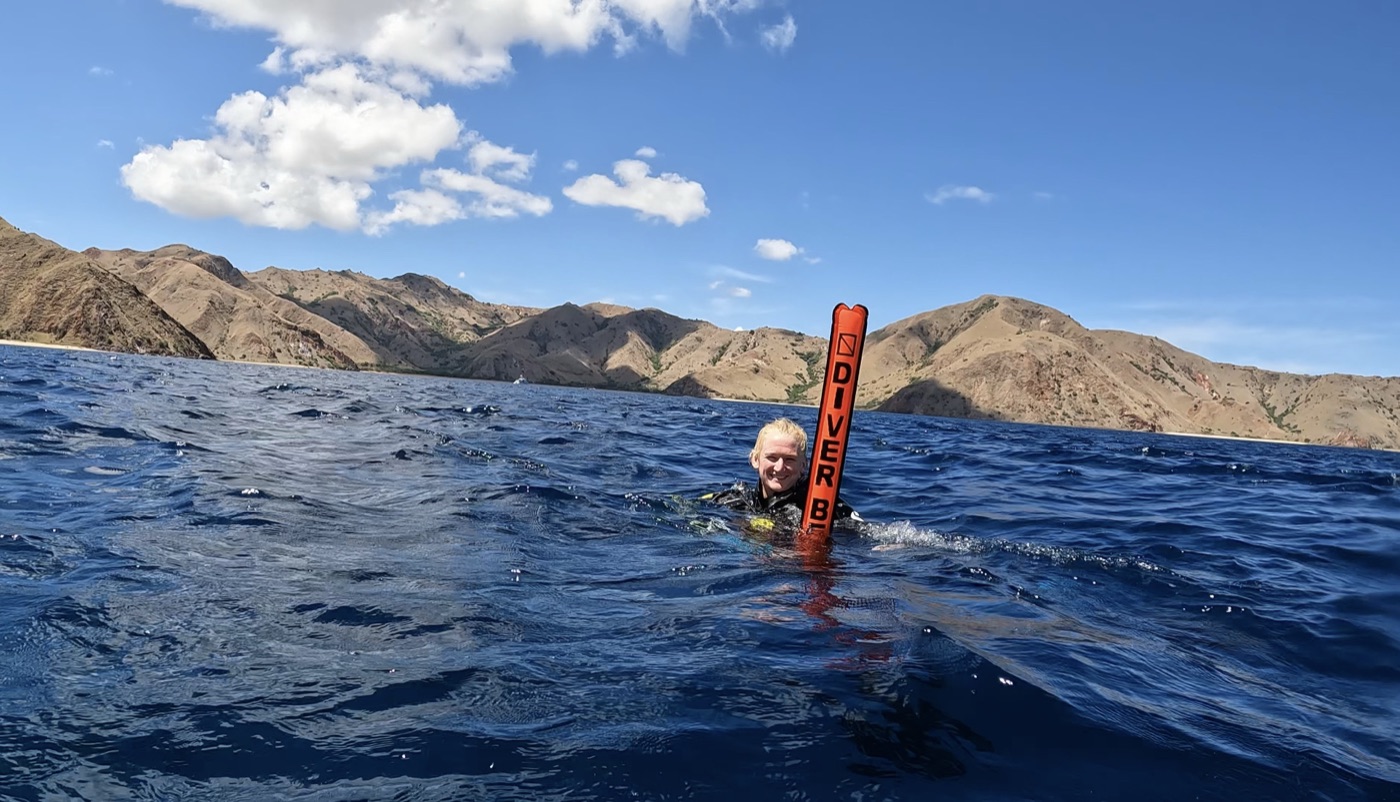 Lucienne surfacing beside a DIVER BELOW marker buoy, with the hills of Komodo behind