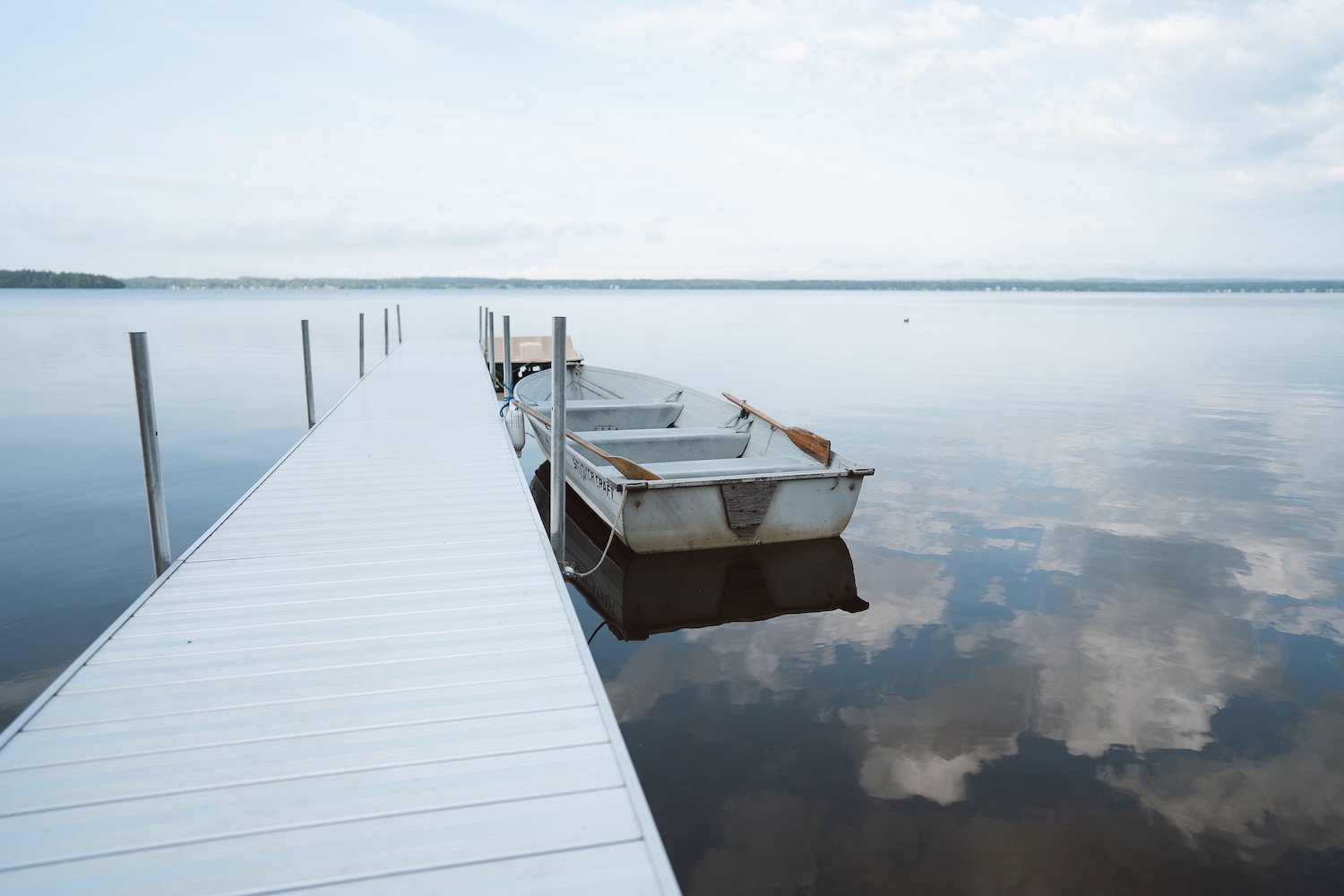 Rowboat tied to a white dock on calm lake water reflecting a cloudy sky.