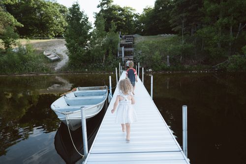 Two children walking barefoot on a dock beside a small rowboat on calm water with trees in the background.