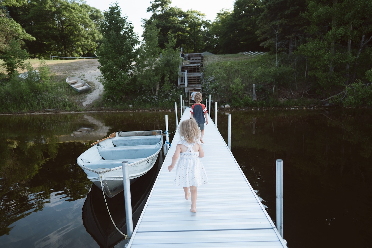 Two children walking barefoot on a dock beside a small rowboat on calm water with trees in the background.