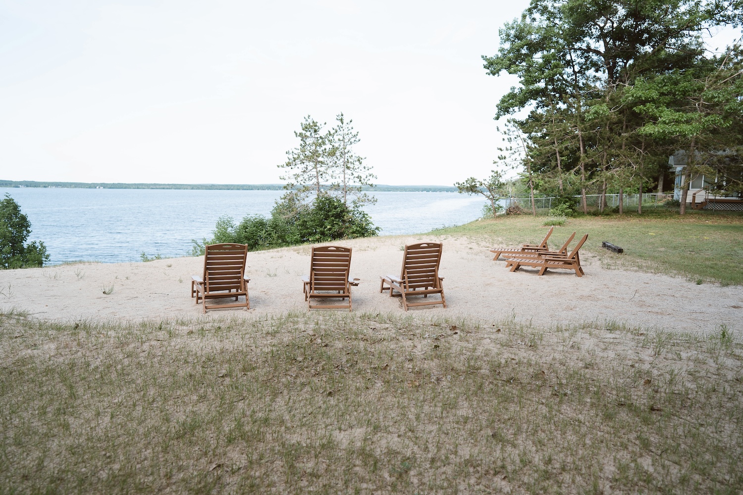 Six wooden lounge chairs arranged on sandy shore facing a large lake with trees in the background.