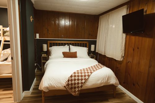 Cozy bedroom with wooden paneled walls, white bed linens, checkered throw, and wall-mounted TV.