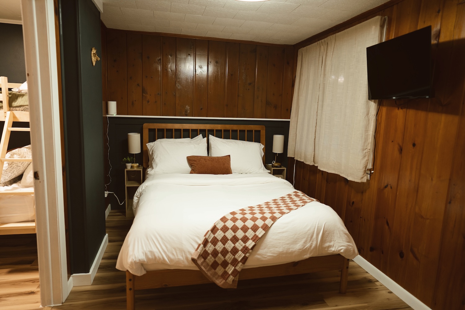 Cozy bedroom with wooden paneled walls, white bed linens, checkered throw, and wall-mounted TV.