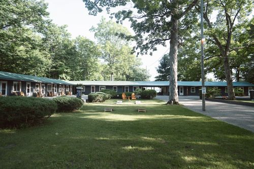 Outdoor view of a motel with green roofs, surrounded by trees and lawns with Adirondack chairs.