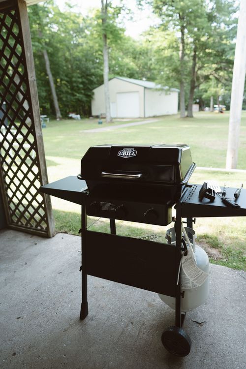 Black Expert Grill propane gas grill on wheels with tools and a propane tank under a porch.