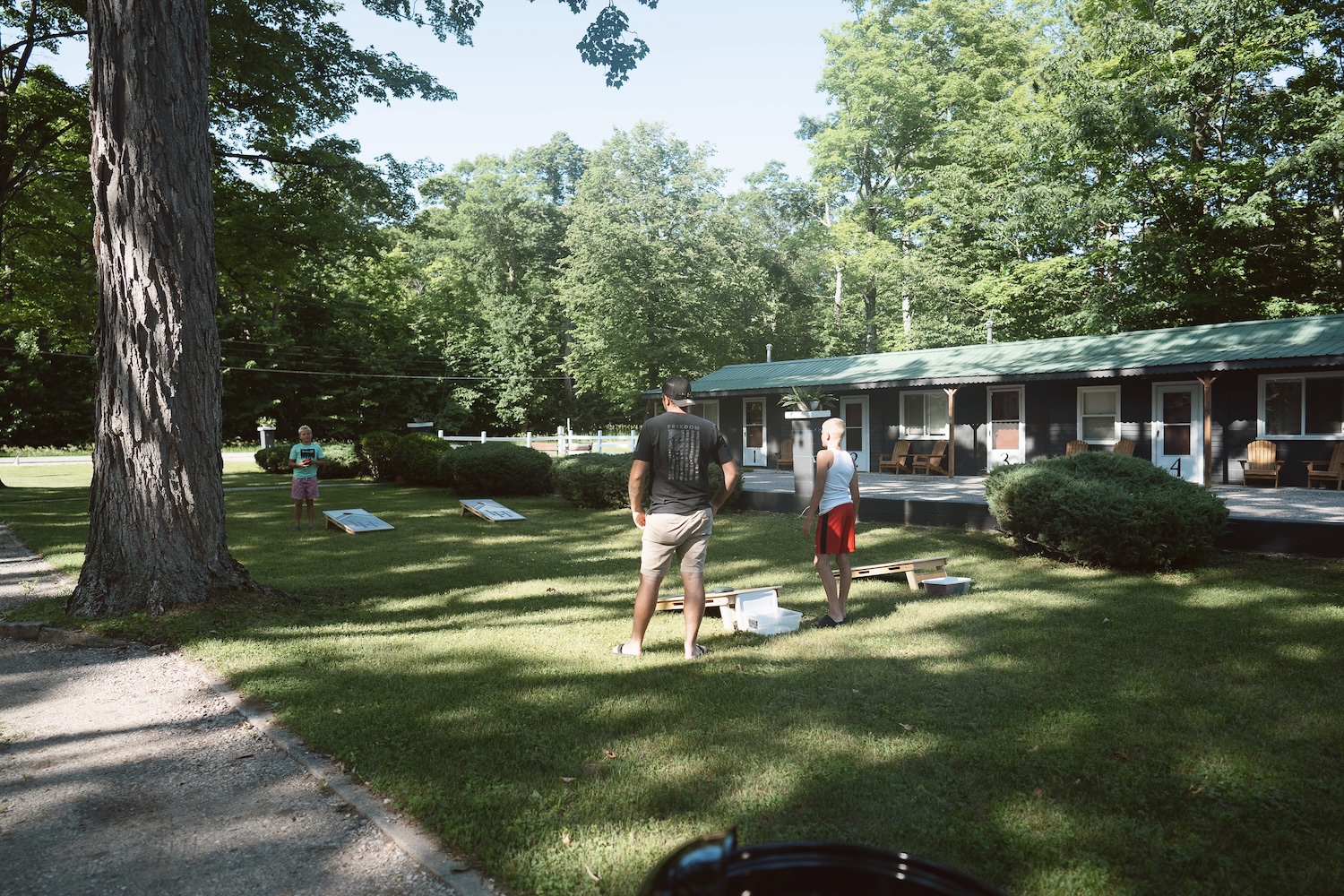 Three people playing cornhole on grass in front of cabins surrounded by trees on a sunny day.