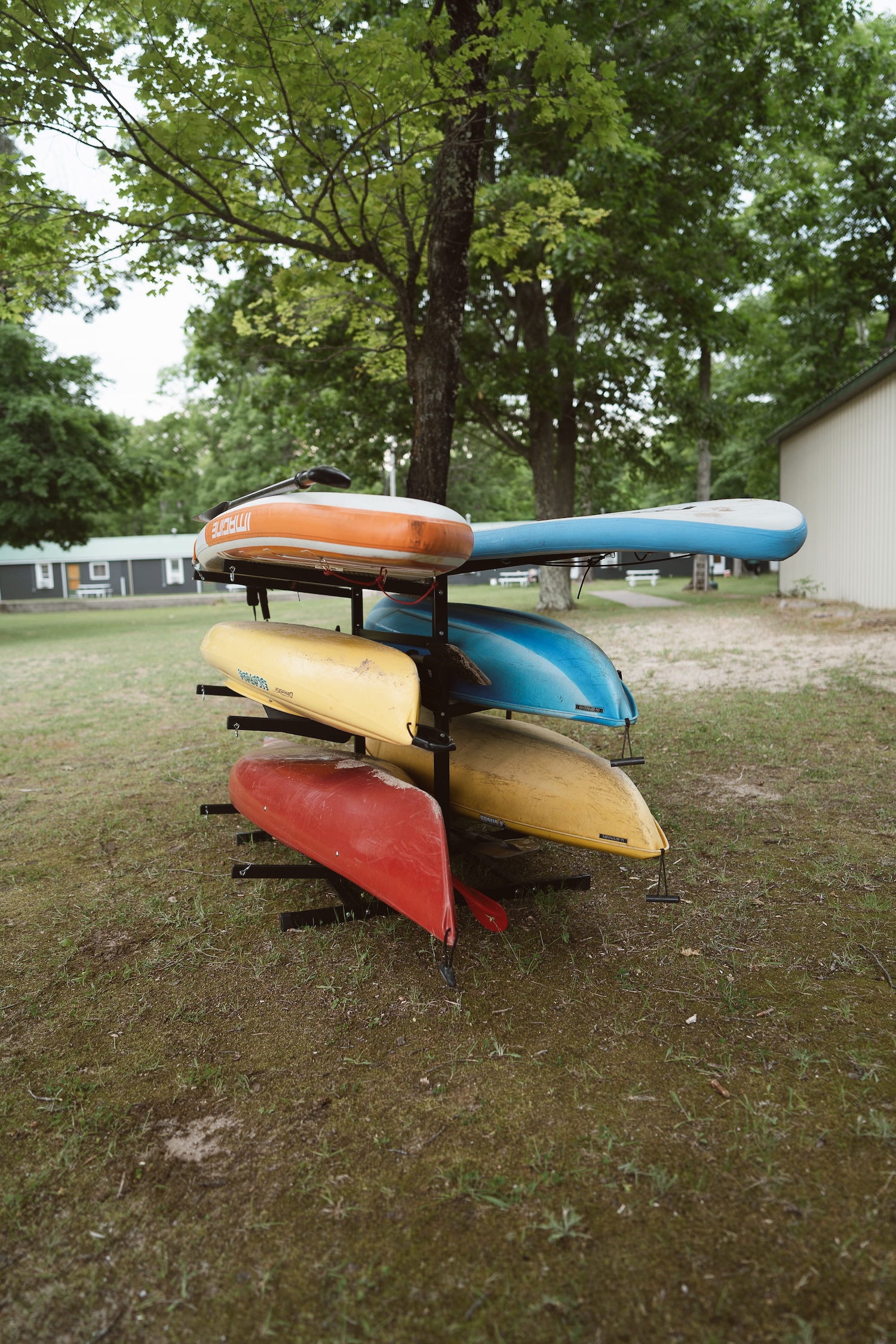 Stack of colorful kayaks and paddleboards on metal racks in a grassy park area with trees.