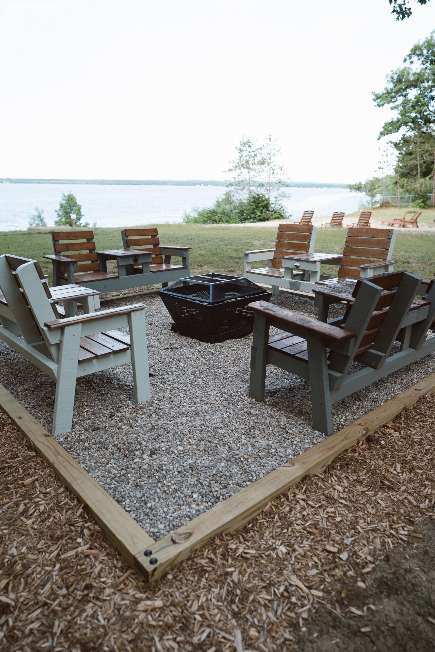 Outdoor fire pit surrounded by wooden chairs and benches on a gravel area near a lake.