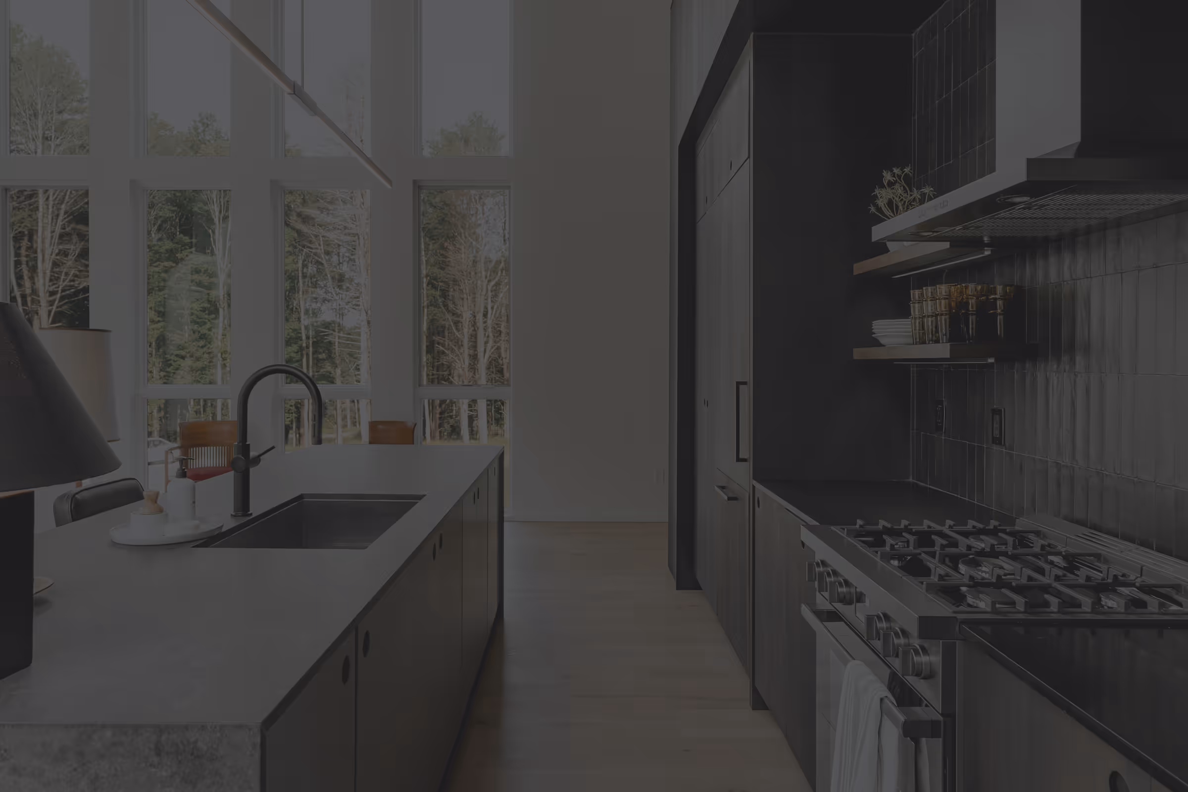 Modern kitchen with black cabinetry, stainless steel gas stove, large island with sink, and tall windows overlooking a wooded area.