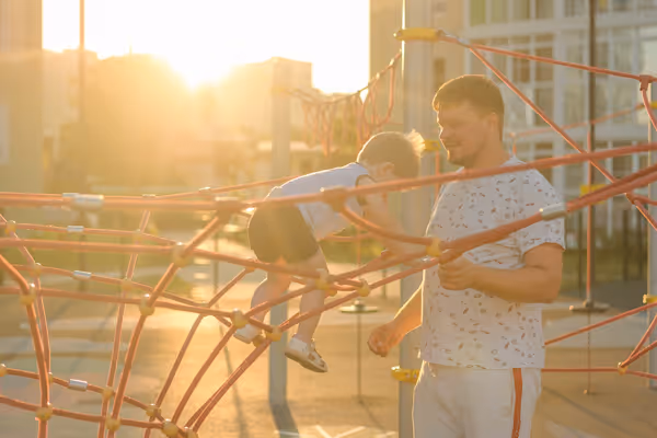Man watching a toddler climb on orange playground climbing ropes at sunset.