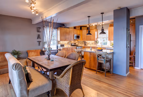 Modern kitchen with wooden cabinets, island seating, wicker and upholstered chairs, and a dining table with decorative sticks in a vase.