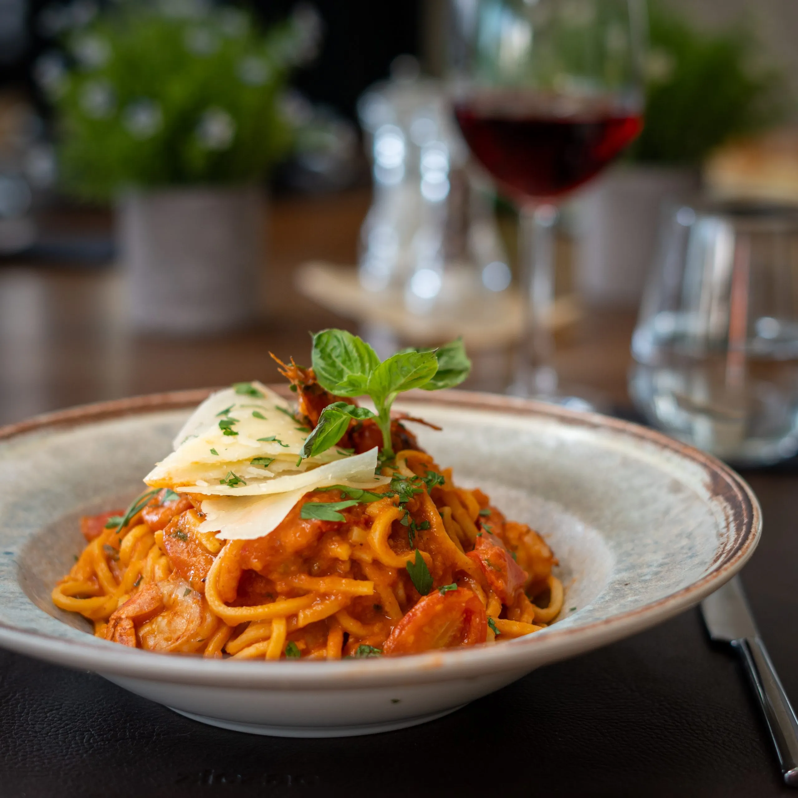 Plate of spaghetti with tomato sauce, shrimp, fresh herbs, and shaved cheese, served on a table with a glass of red wine in the background.