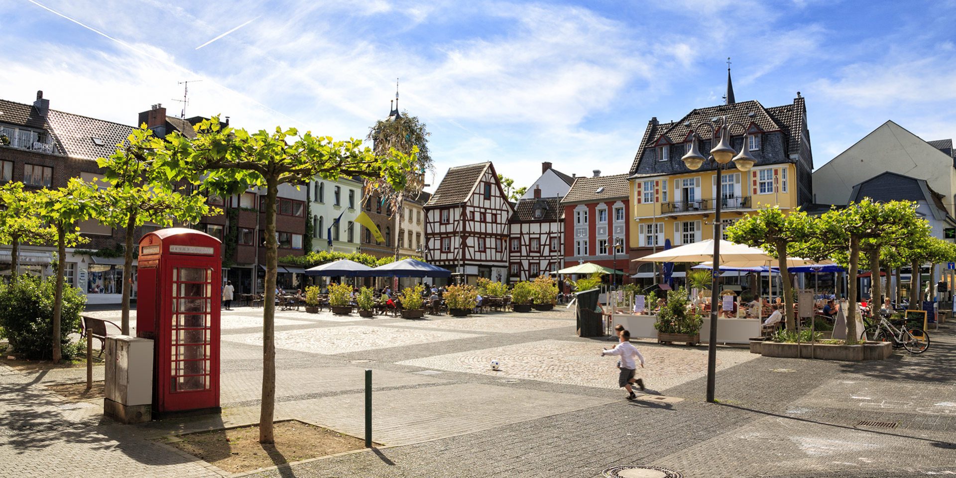 Stadtplatz mit Fachwerkhäusern, roten Telefonzelle, Cafés und Kind beim Fußballspielen.