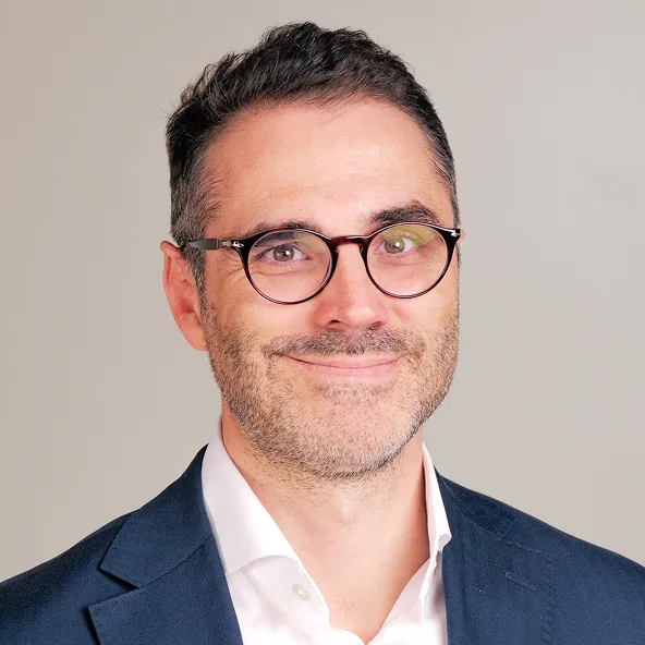 Portrait d'un homme souriant portant des lunettes rondes, une chemise blanche et une veste bleu foncé, avec un fond neutre.