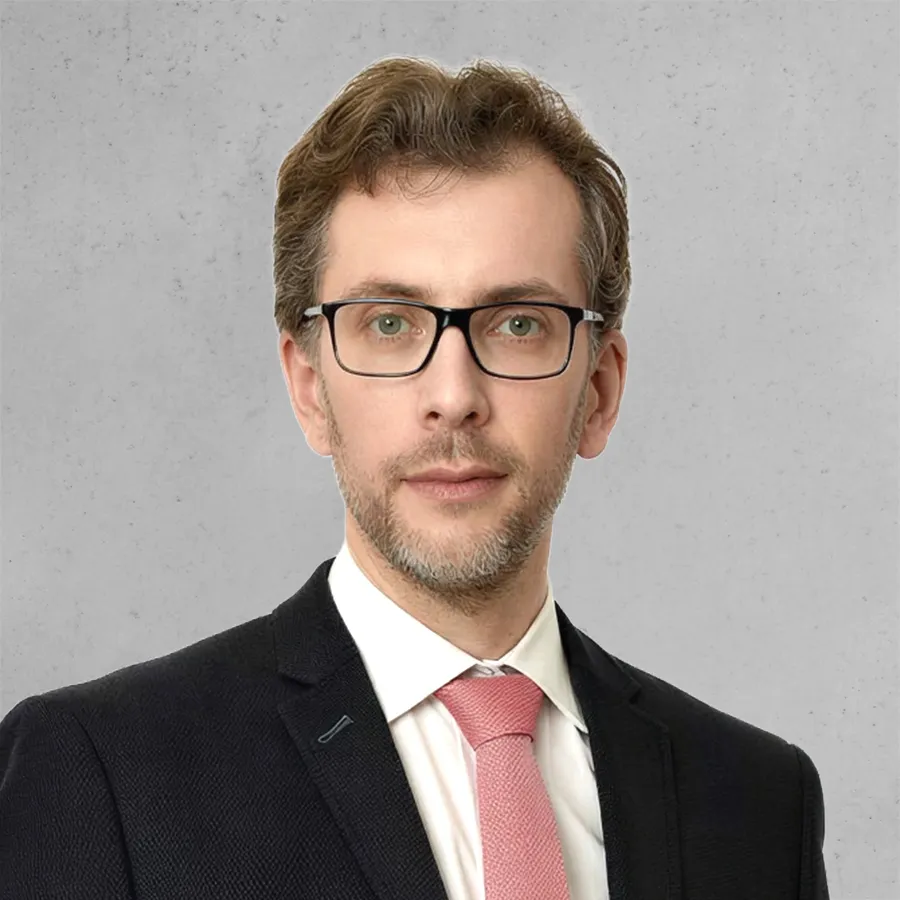 Homme portant un costume noir, une chemise blanche et une cravate rose, avec des lunettes et une barbe courte.