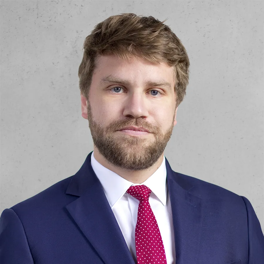 Portrait d'un homme aux cheveux bruns et barbe, vêtu d'un costume bleu marine et d'une chemise blanche, fond gris clair.
