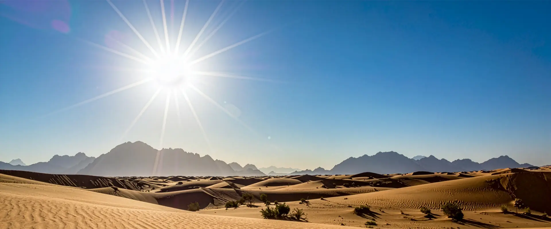 Bright sun shining over a vast desert with sand dunes and distant mountain range under a clear blue sky.