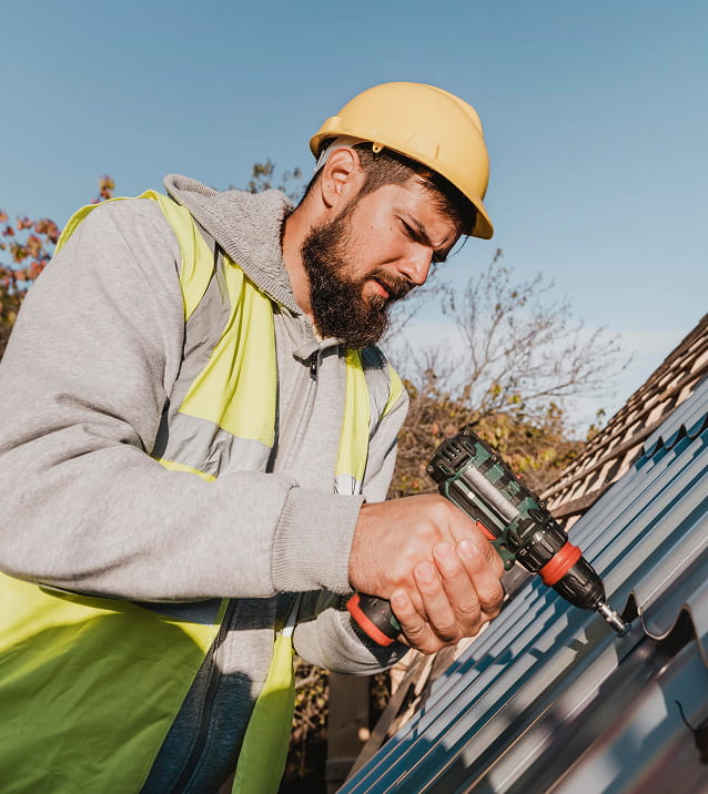 Bearded construction worker wearing a yellow hard hat and safety vest using a power drill on a metal roof under clear blue sky.