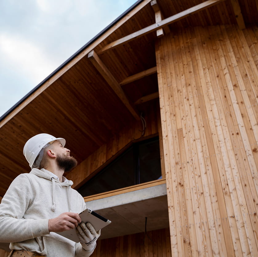 Construction worker in a white helmet and hoodie holding a tablet, inspecting a wooden building exterior.