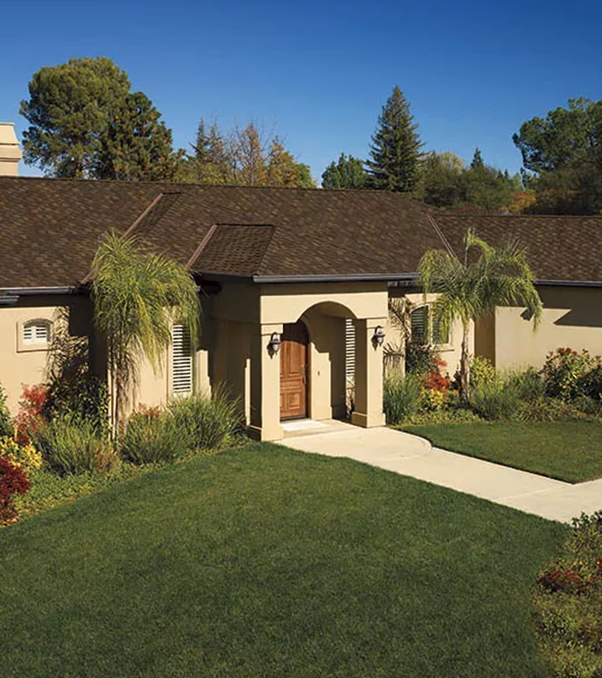 Single-story beige house with a brown shingle roof, wooden front door under an archway, surrounded by green lawn and palm trees.