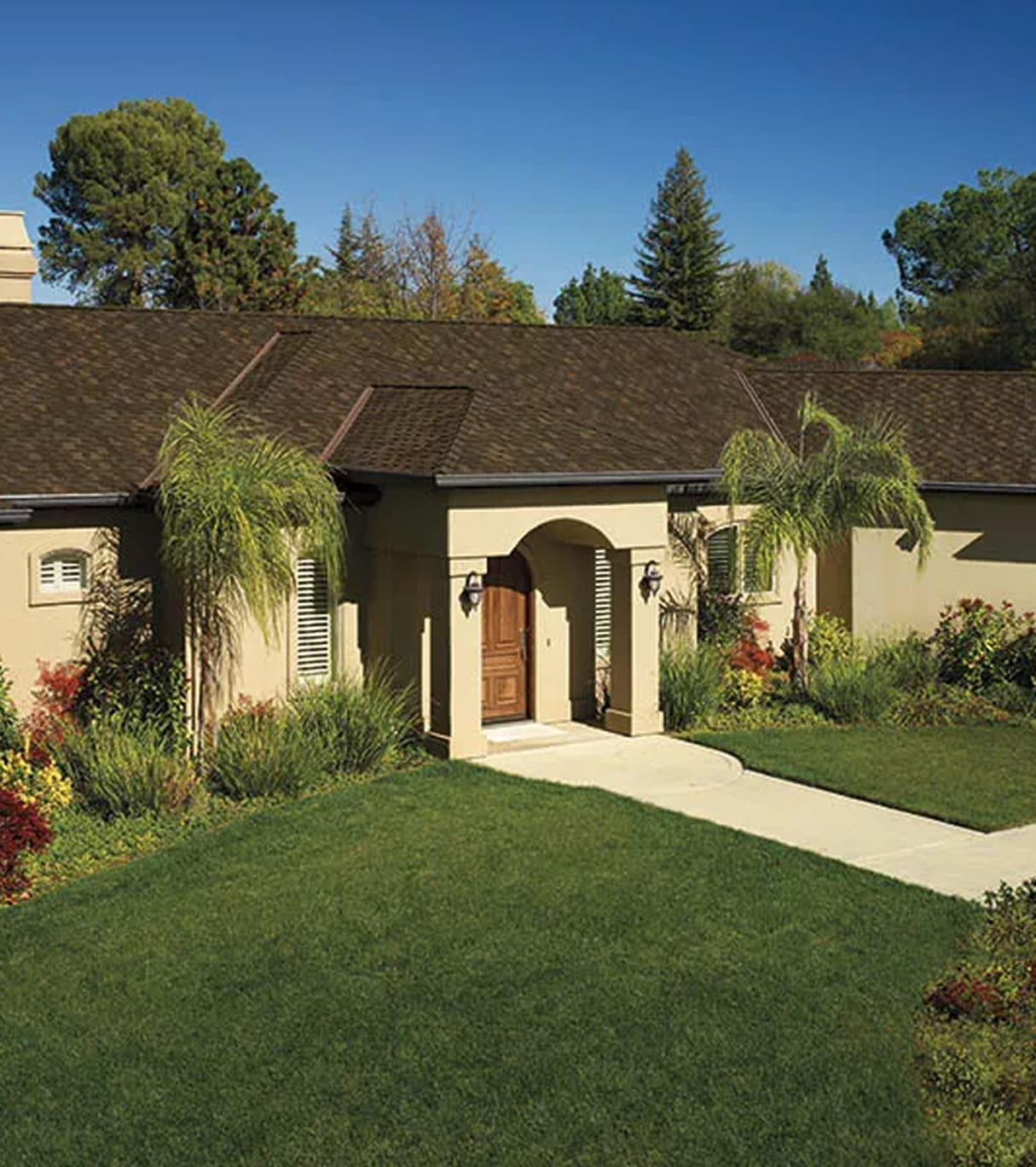 Single-story beige house with a brown shingle roof, wooden front door under an archway, surrounded by green lawn and palm trees.