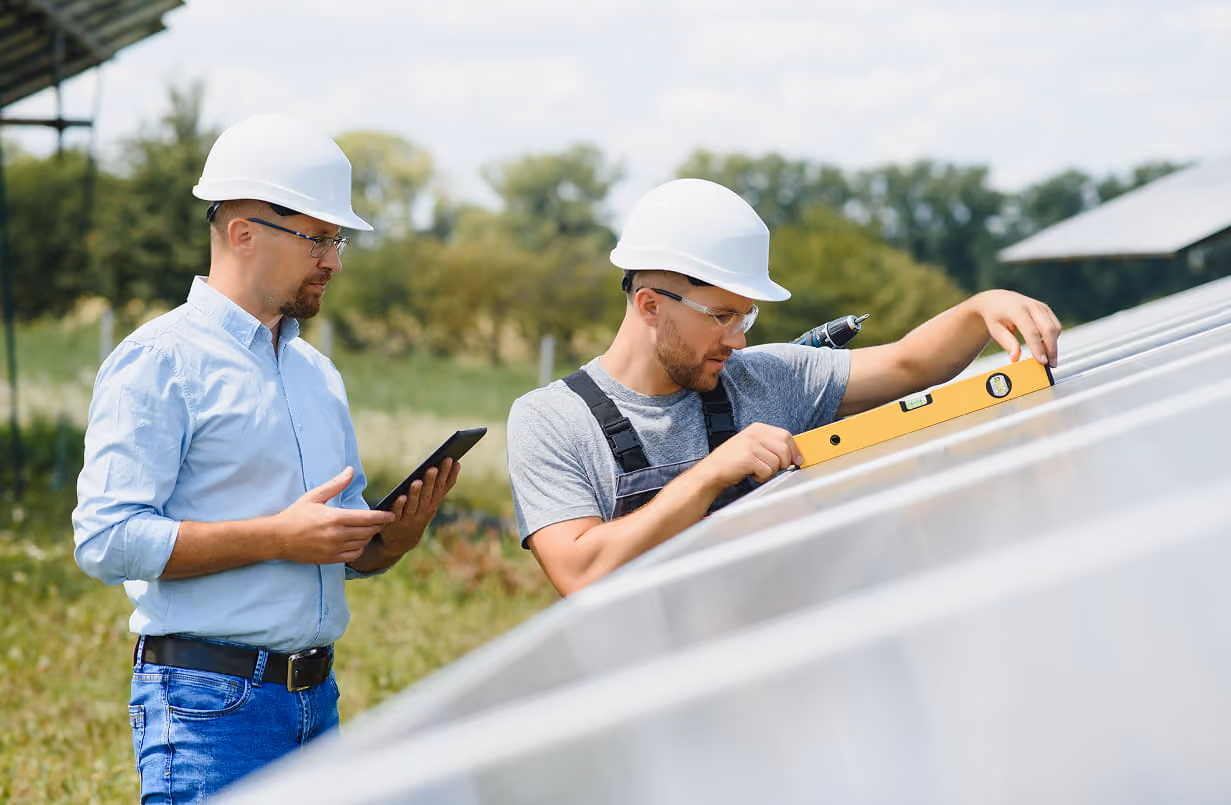 Two men in hard hats inspecting solar panels outdoors, one using a spirit level and the other holding a tablet.