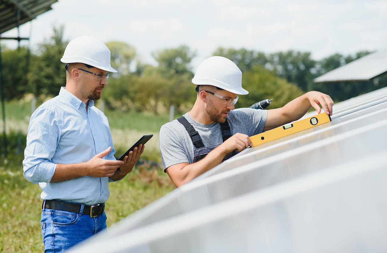 Two men in hard hats inspecting solar panels outdoors, one using a spirit level and the other holding a tablet.