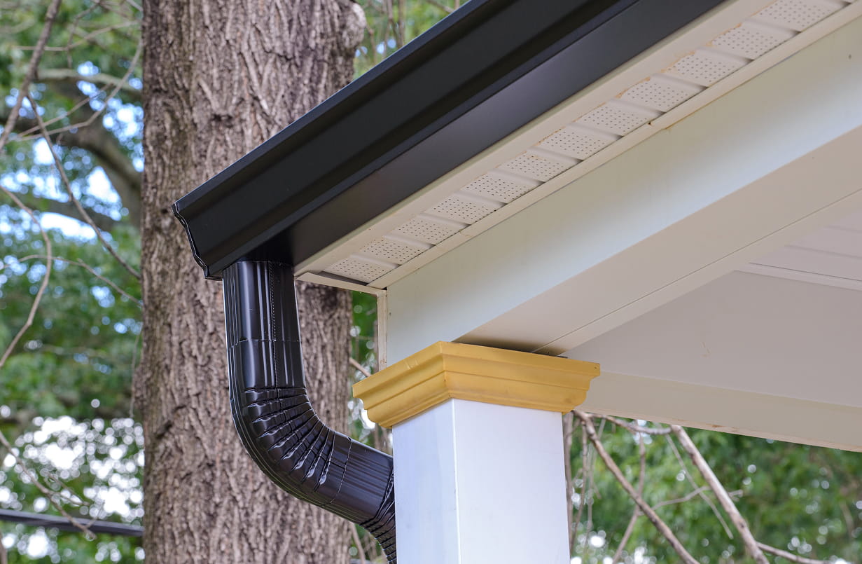 Close-up of a black metal gutter and downspout attached to the corner of a white porch roof with a yellow trim and tree trunk in the background.