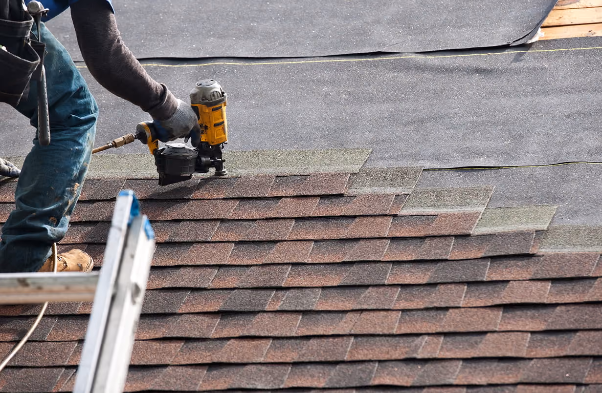 Roofer installing brown asphalt shingles on a roof using a nail gun with underlayment visible underneath.