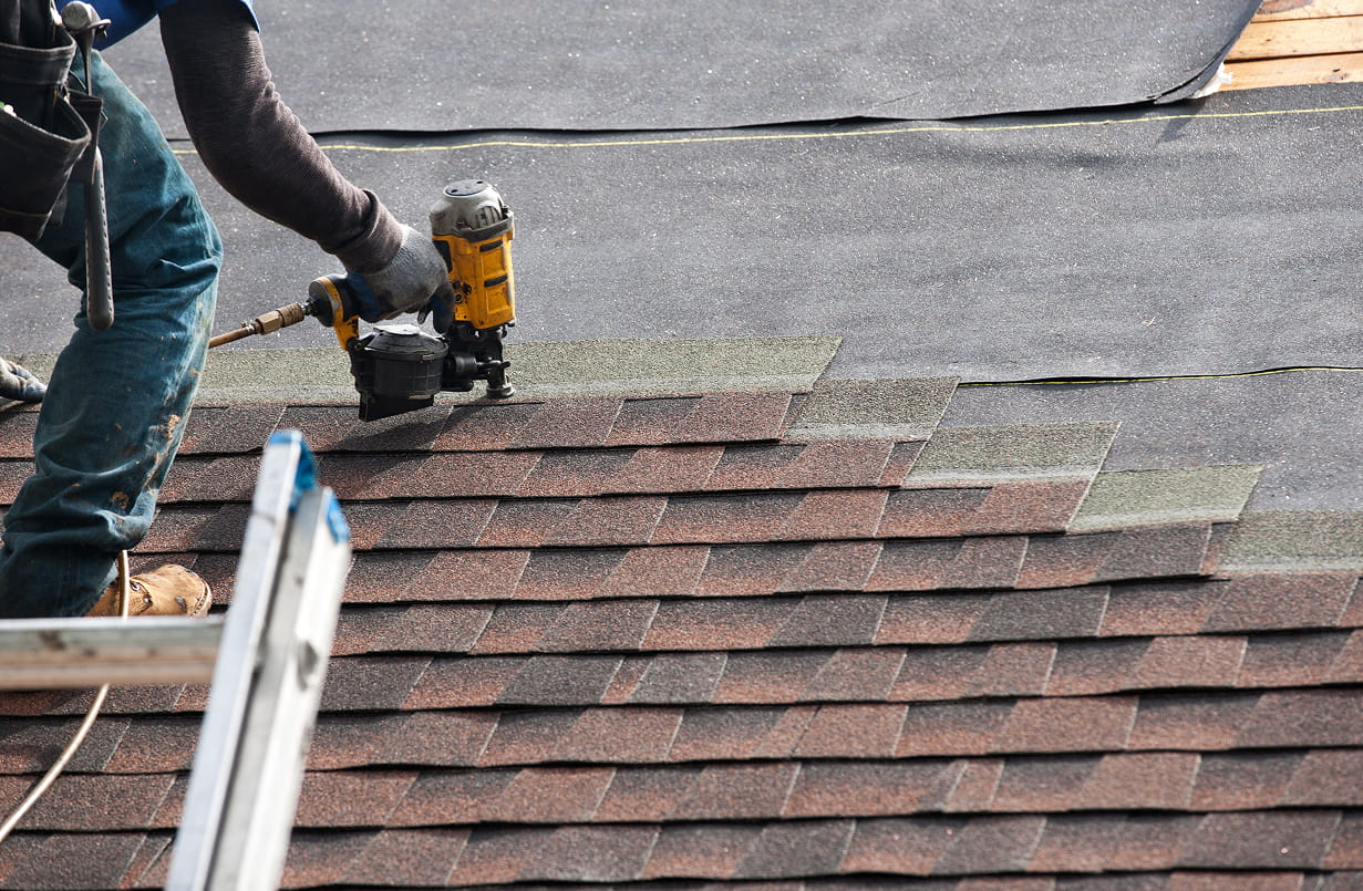 Roofer installing brown asphalt shingles on a roof using a nail gun with underlayment visible underneath.