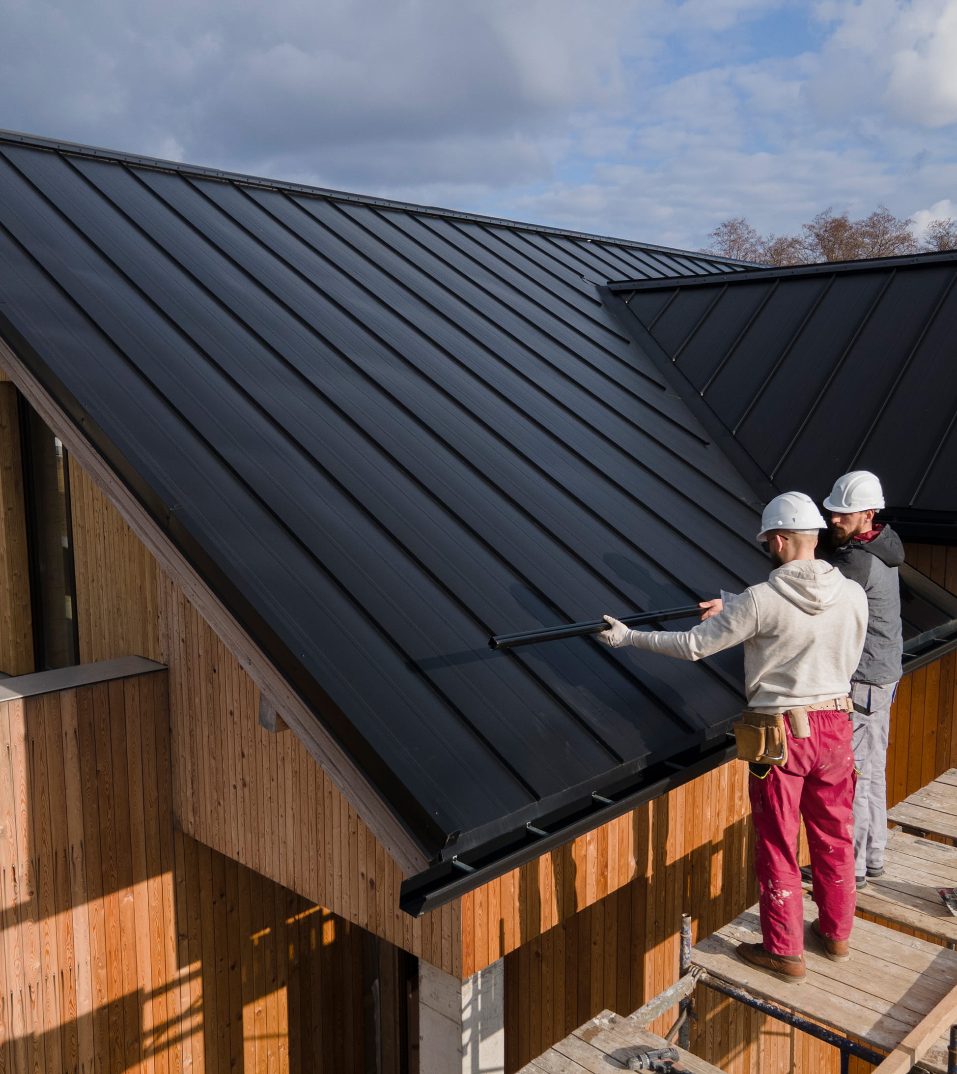 Two construction workers wearing helmets installing a black metal roof on a wooden building.