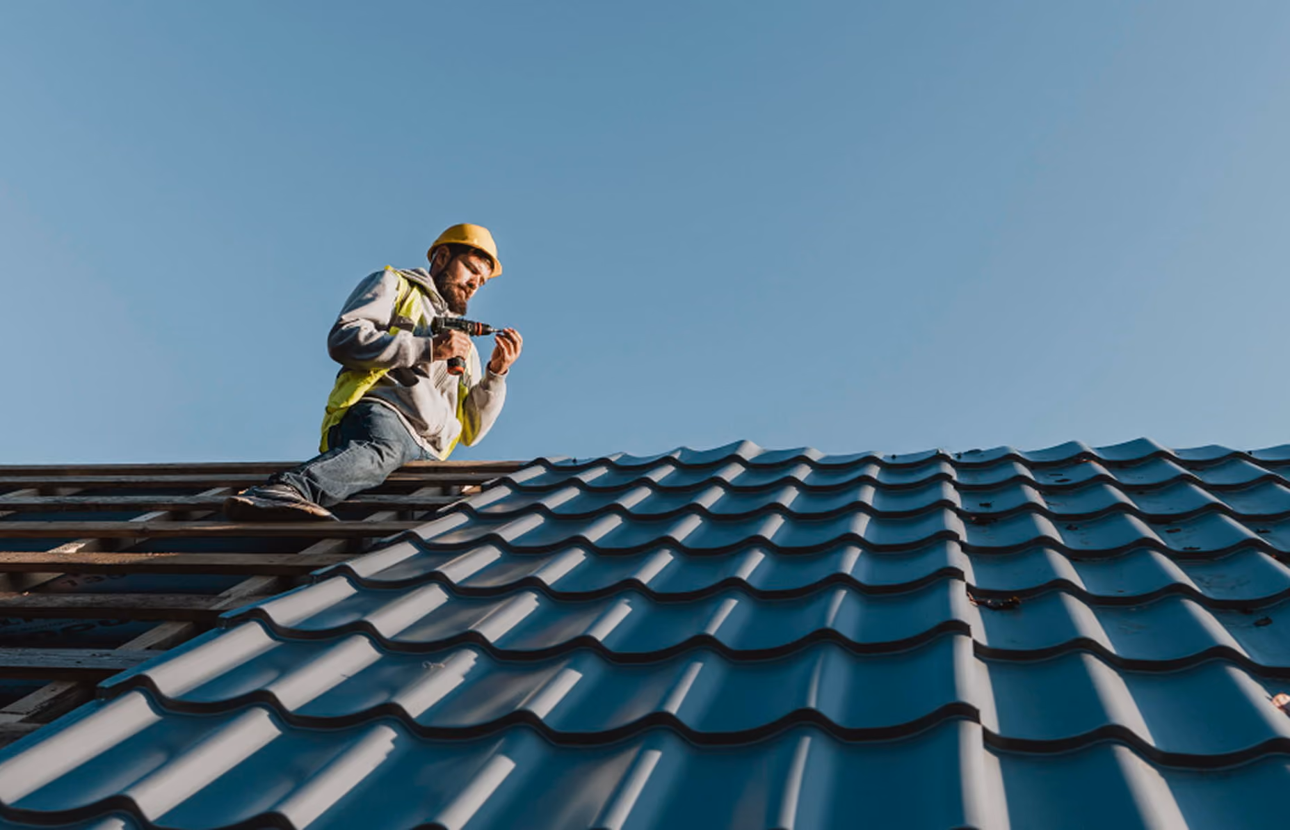 Construction worker in a yellow safety helmet and vest installing metal roofing panels on a clear day.