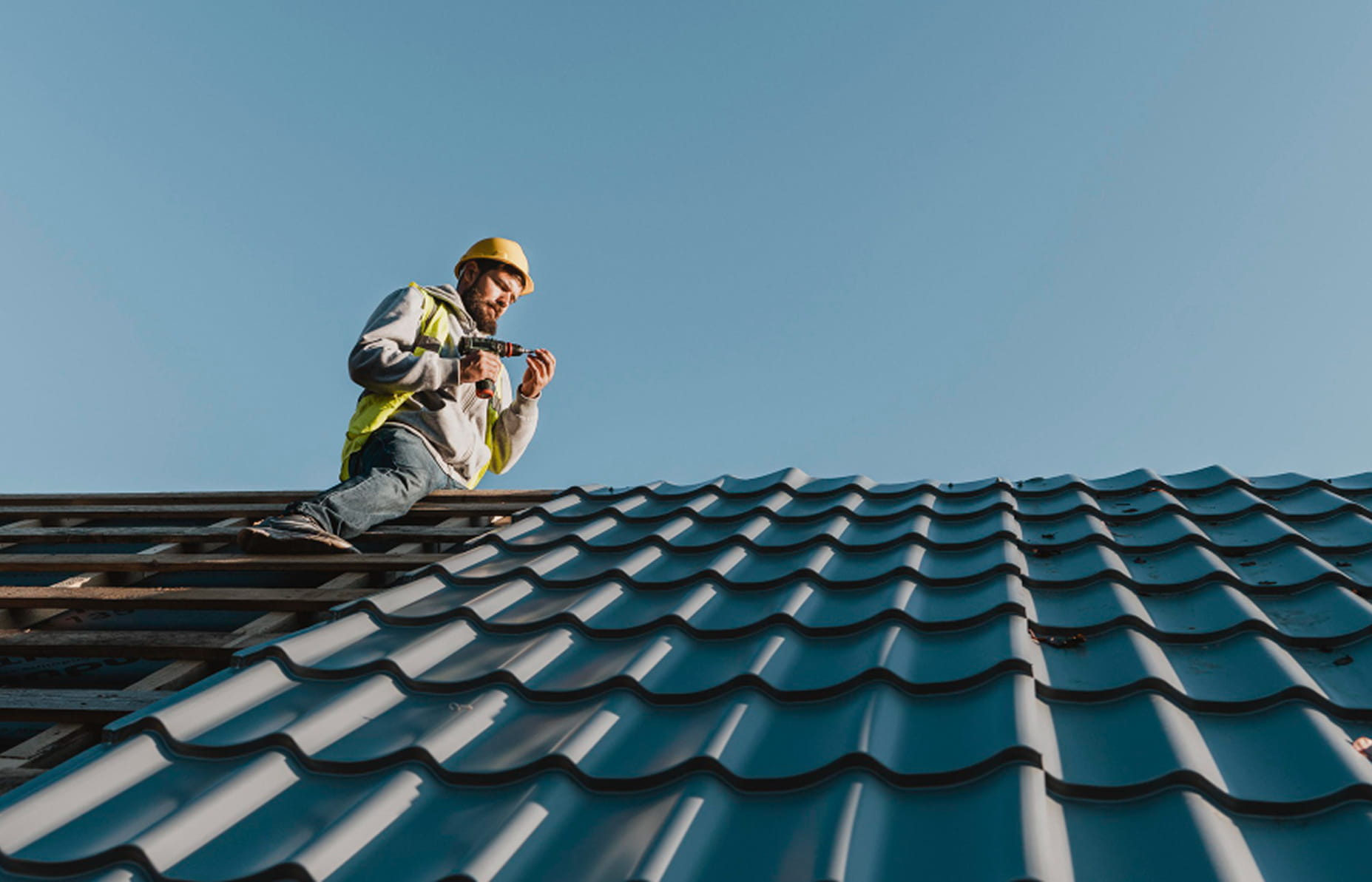 Construction worker in a yellow safety helmet and vest installing metal roofing panels on a clear day.