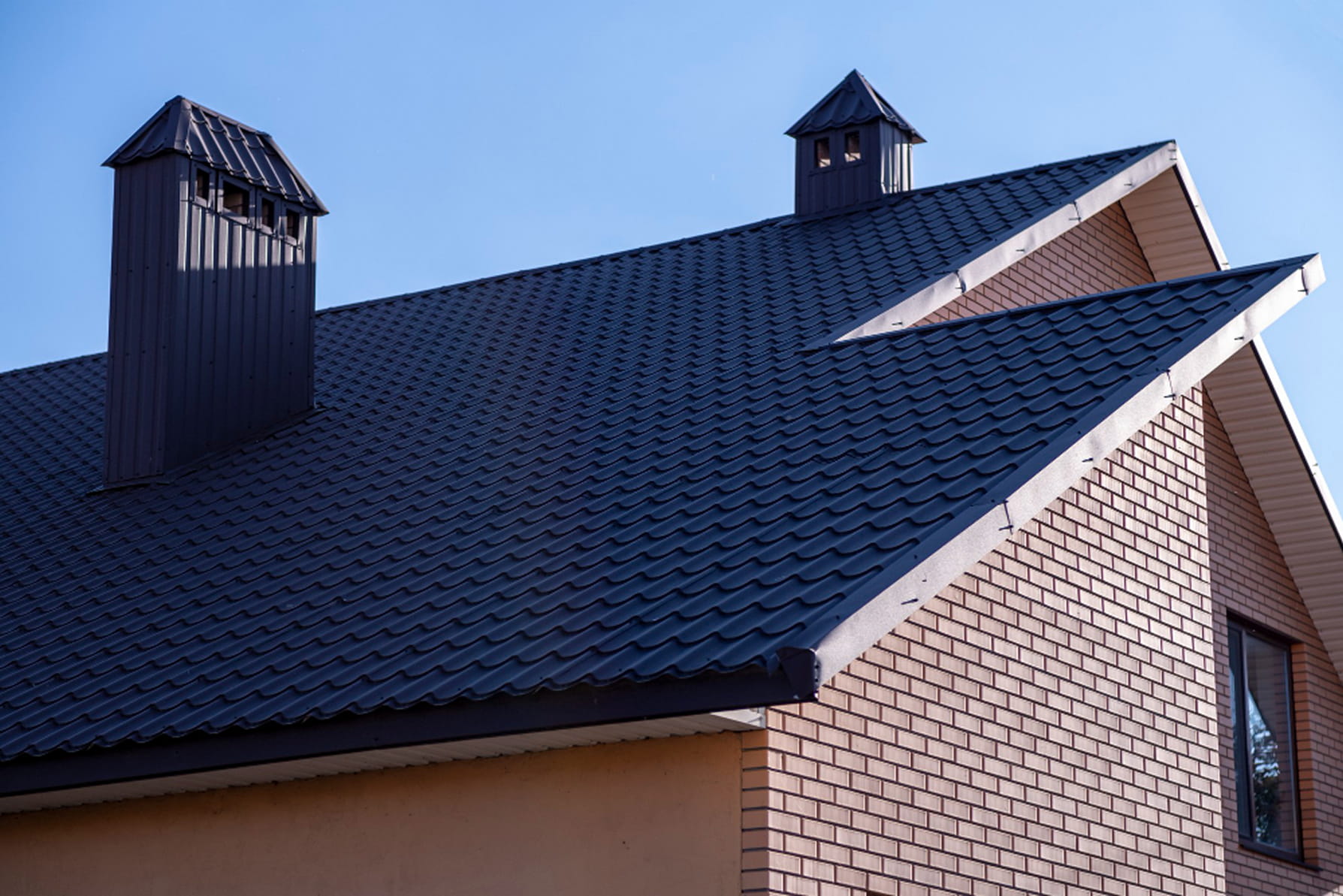 House roof with dark blue metal tiles and two small metal chimneys under clear blue sky.