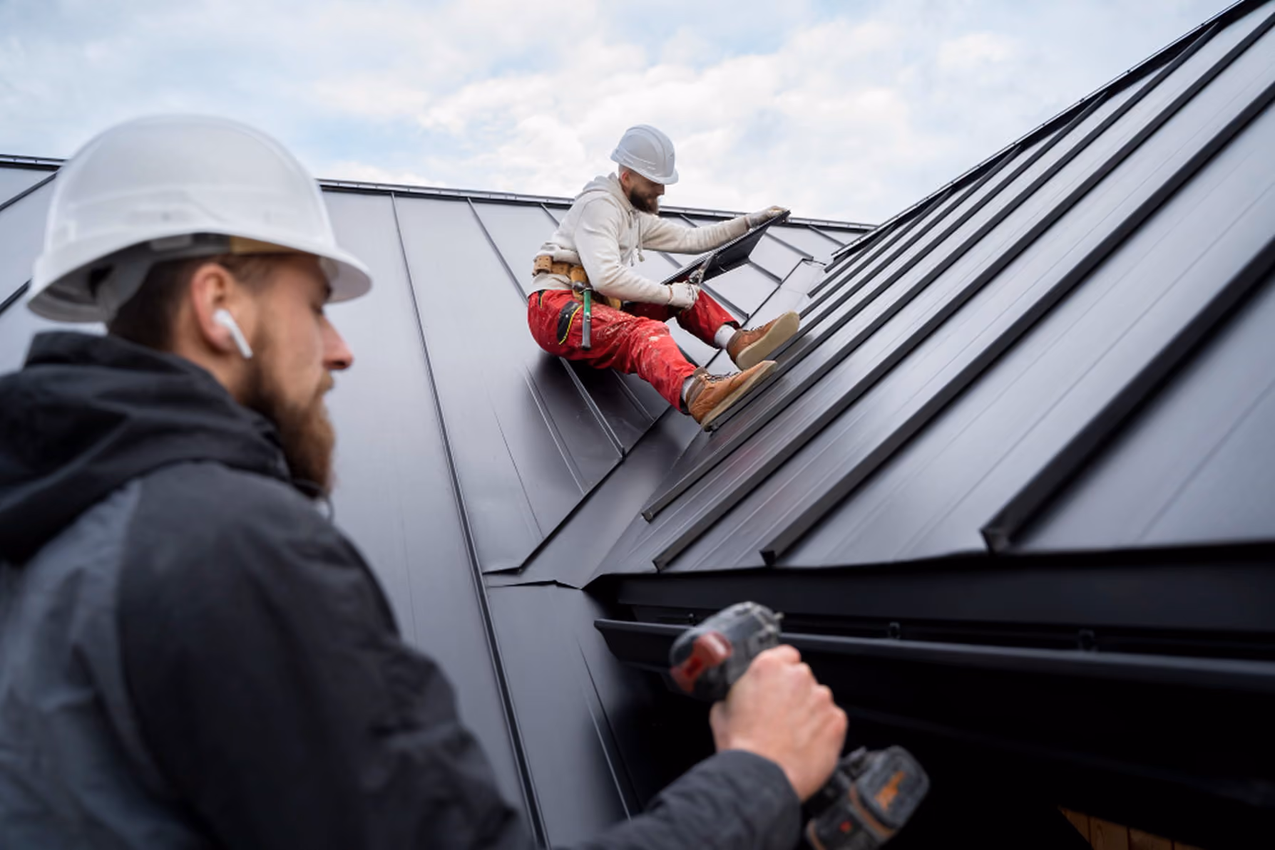 Two construction workers wearing helmets installing metal roofing panels on a steep roof.