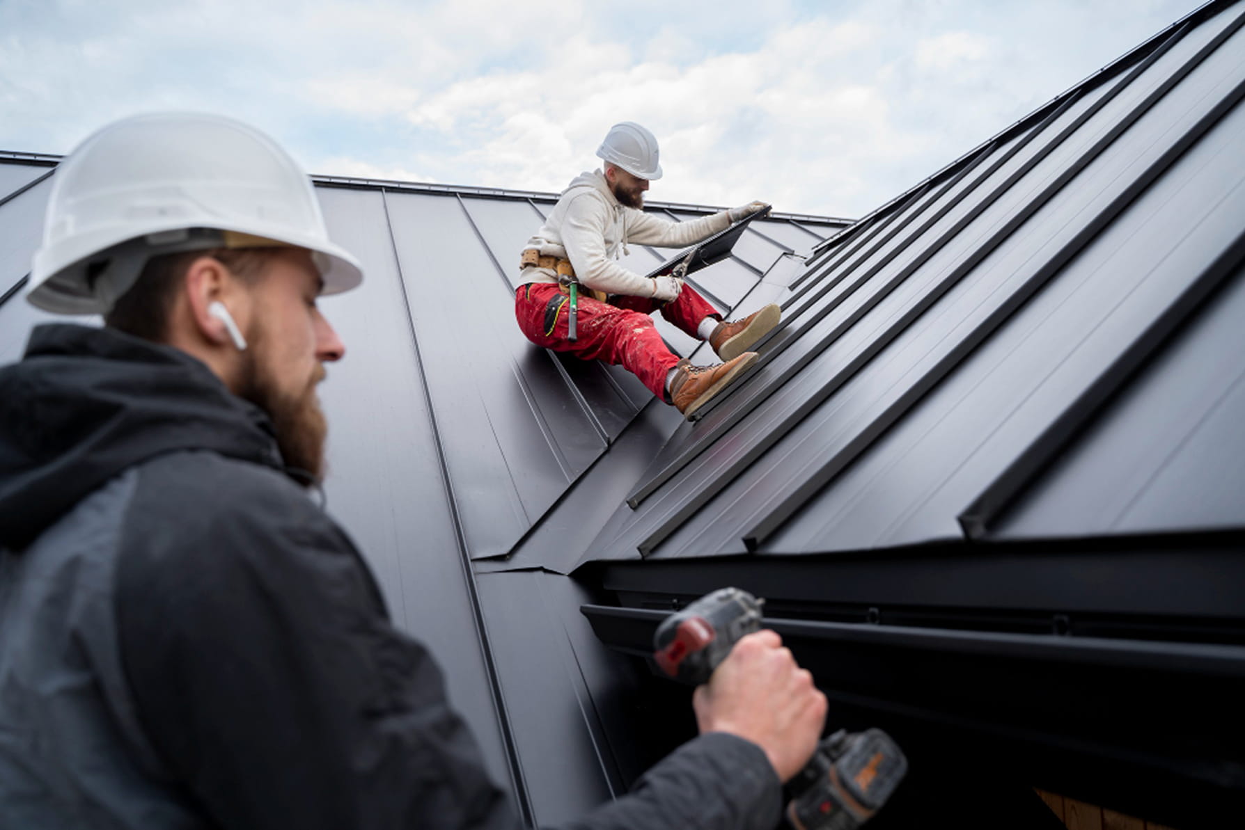 Two construction workers wearing helmets installing metal roofing panels on a steep roof.