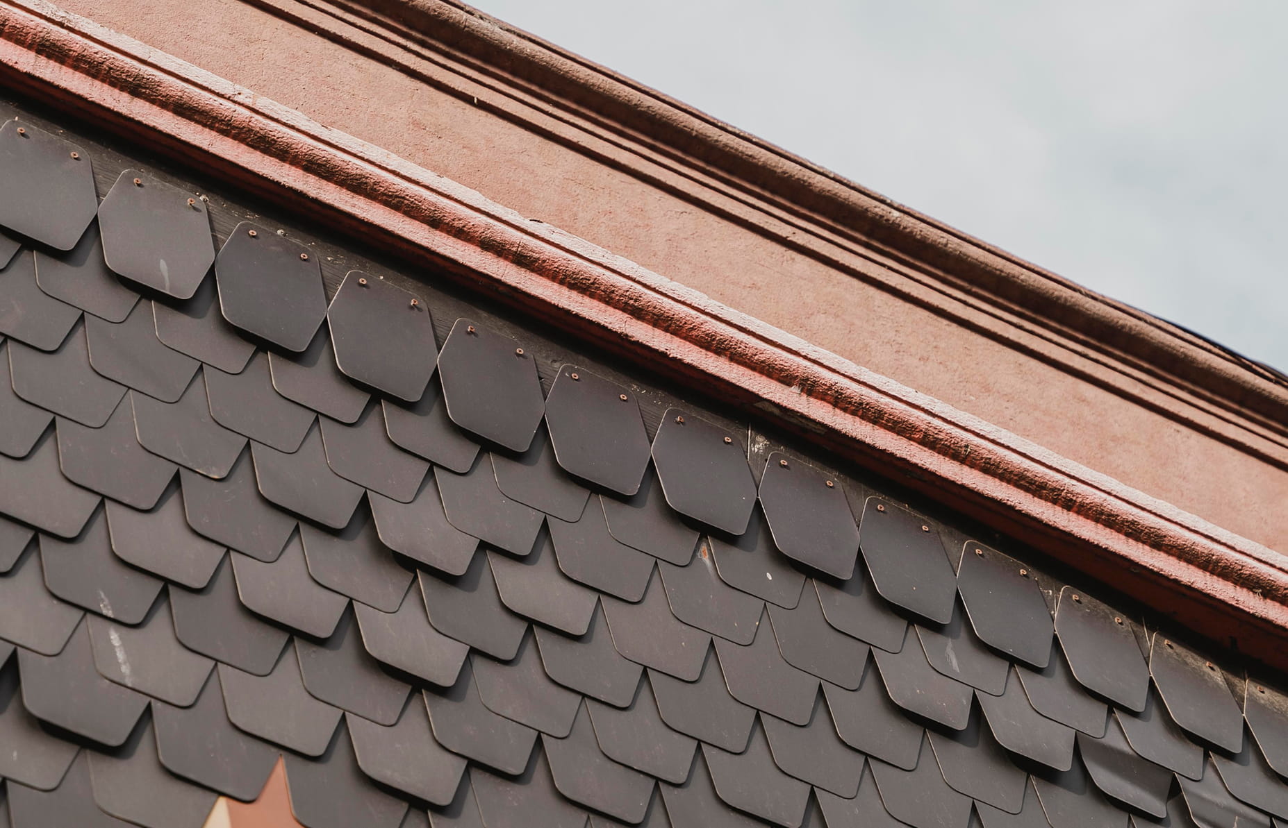 Close-up of a roof with overlapping dark gray shingles beneath a reddish-brown brick or stone ledge.