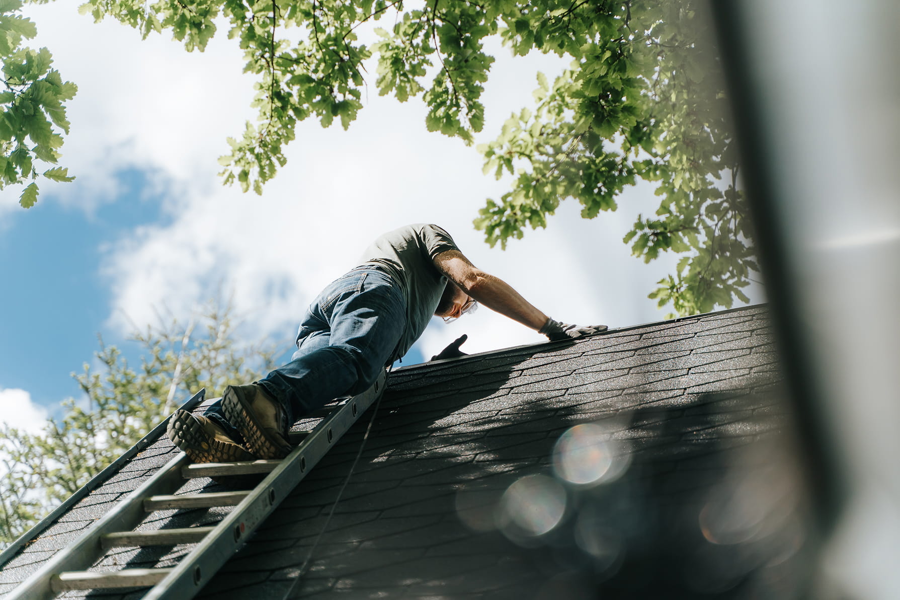 Man wearing gloves climbing a ladder to work on a roof under a blue sky with tree branches overhead.