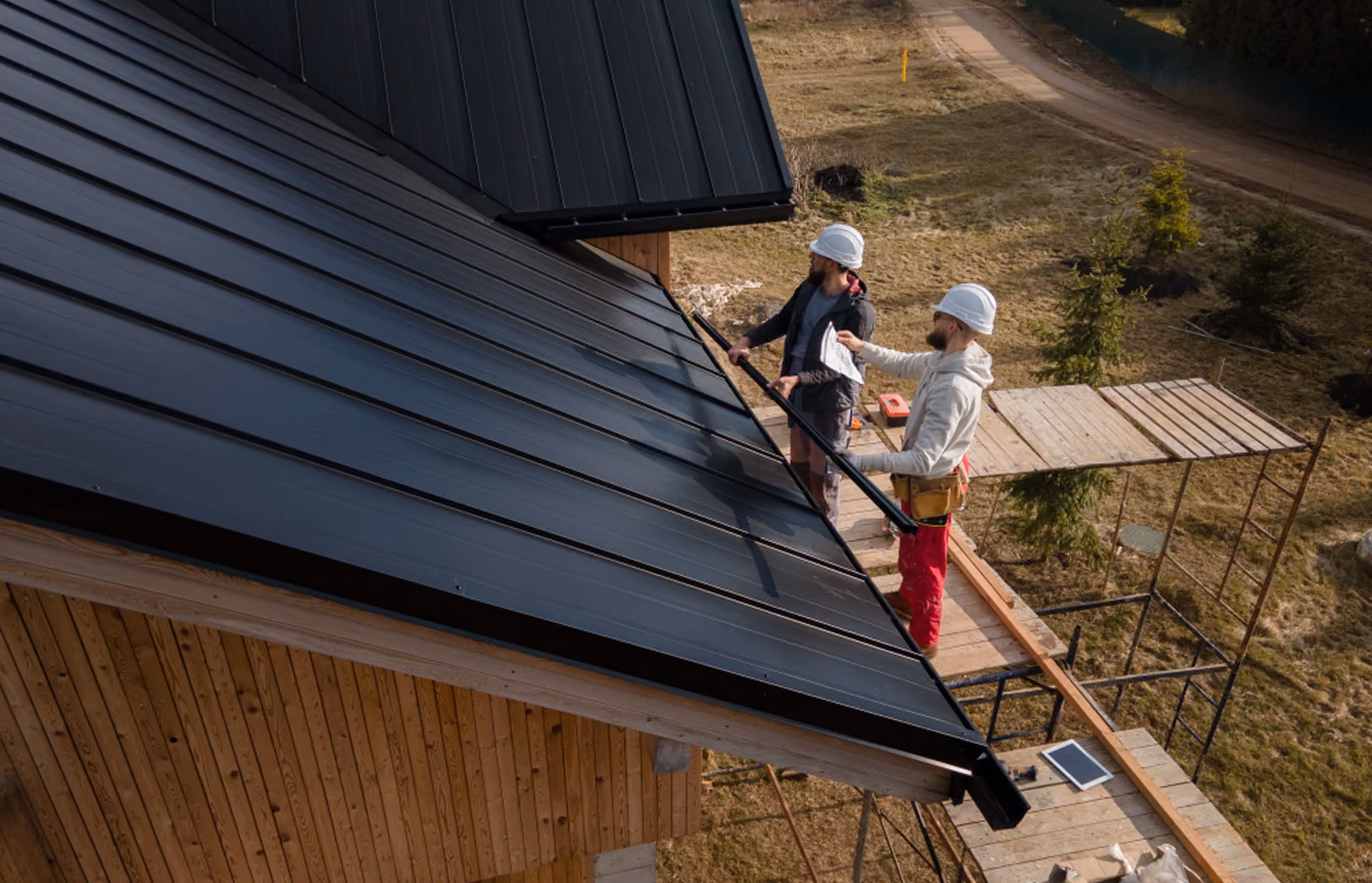 Two construction workers wearing white helmets installing a black metal roof on a wooden house.