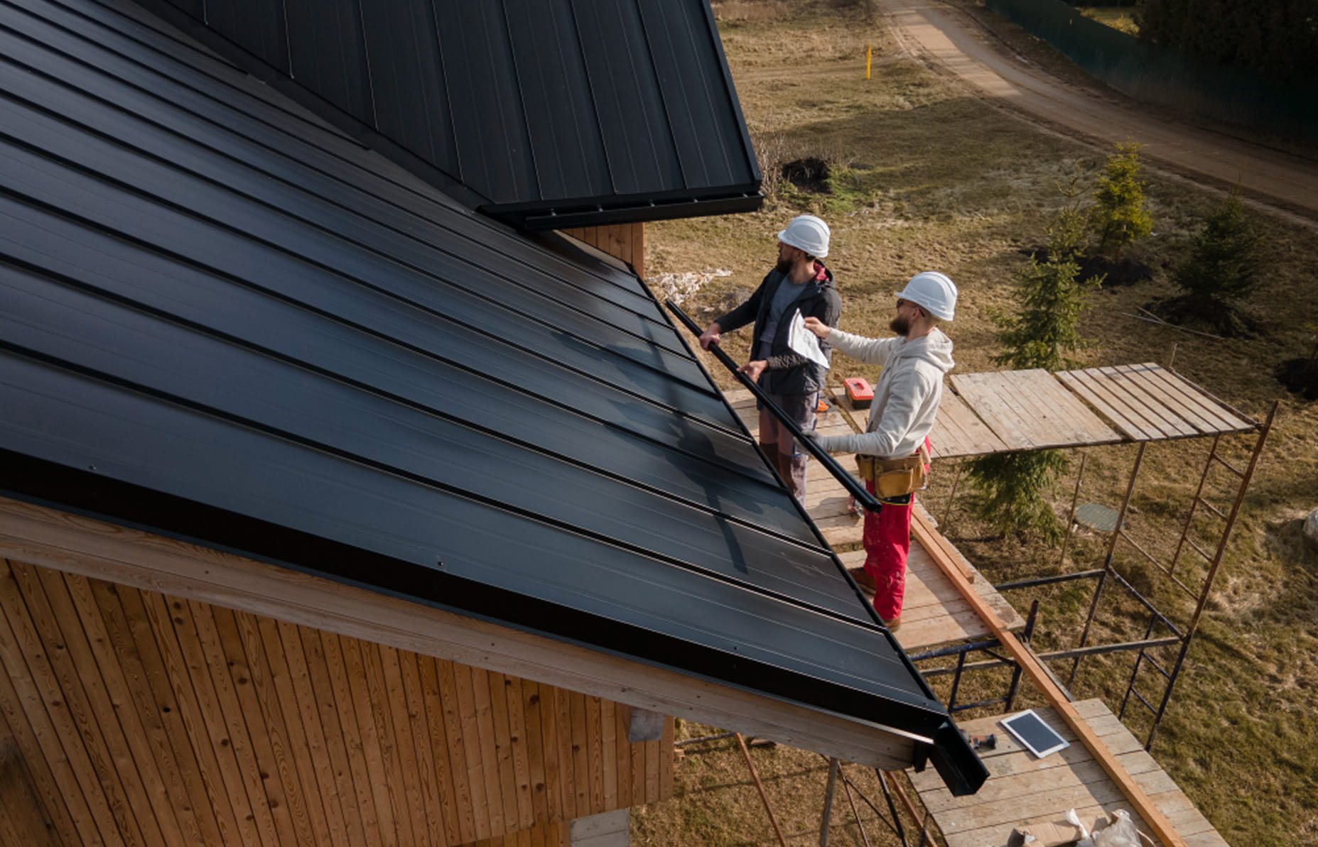 Two construction workers wearing white helmets installing a black metal roof on a wooden house.