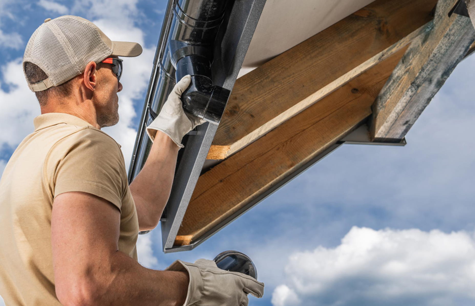 Man wearing gloves and cap installing a black gutter pipe on a wooden roof edge under a partly cloudy sky.