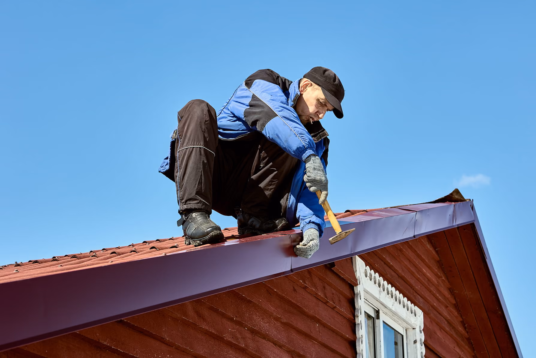 Man in blue jacket and black cap working on a red roof using a hammer under clear blue sky.