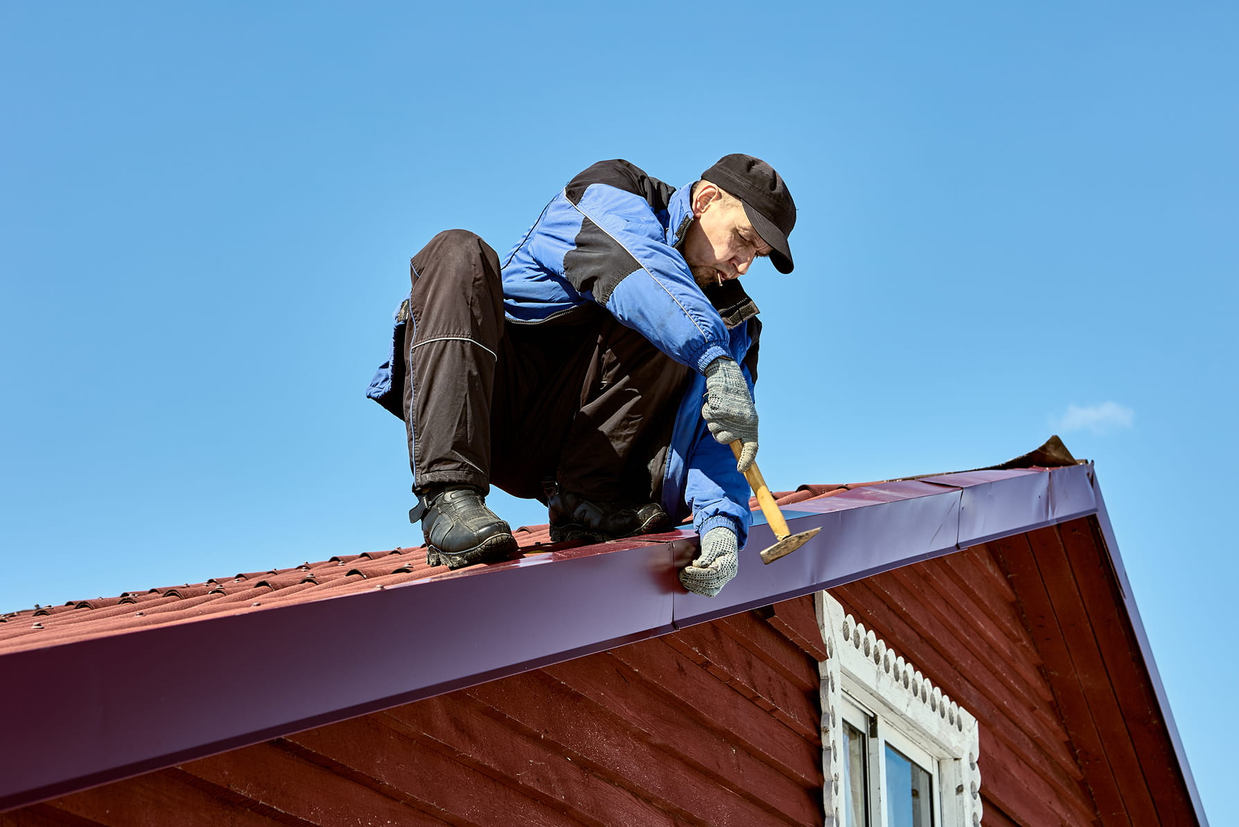 Man in blue jacket and black cap working on a red roof using a hammer under clear blue sky.