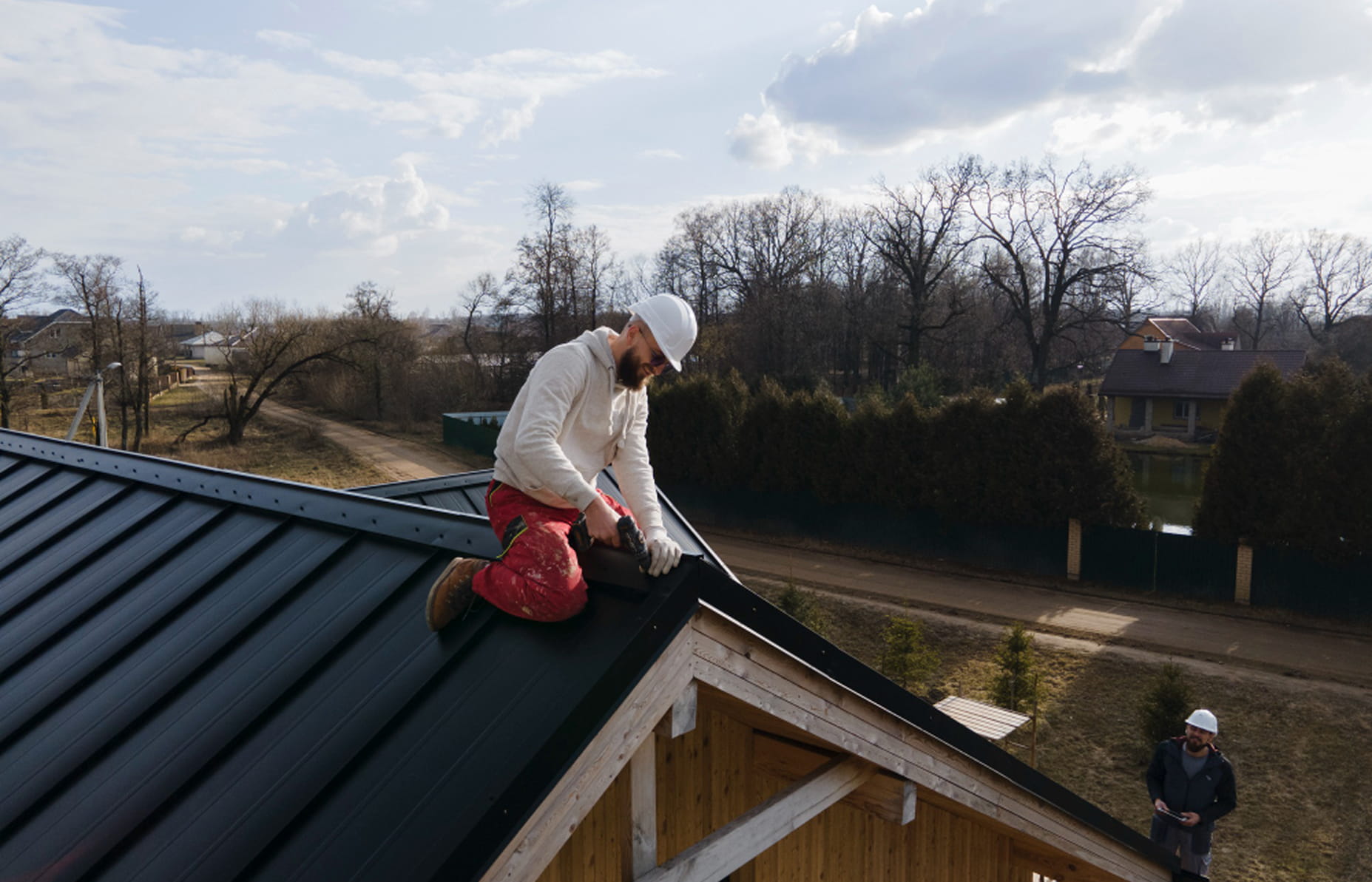Construction worker wearing a white helmet and red pants kneeling on a black metal roof, using a power tool to install roofing material.